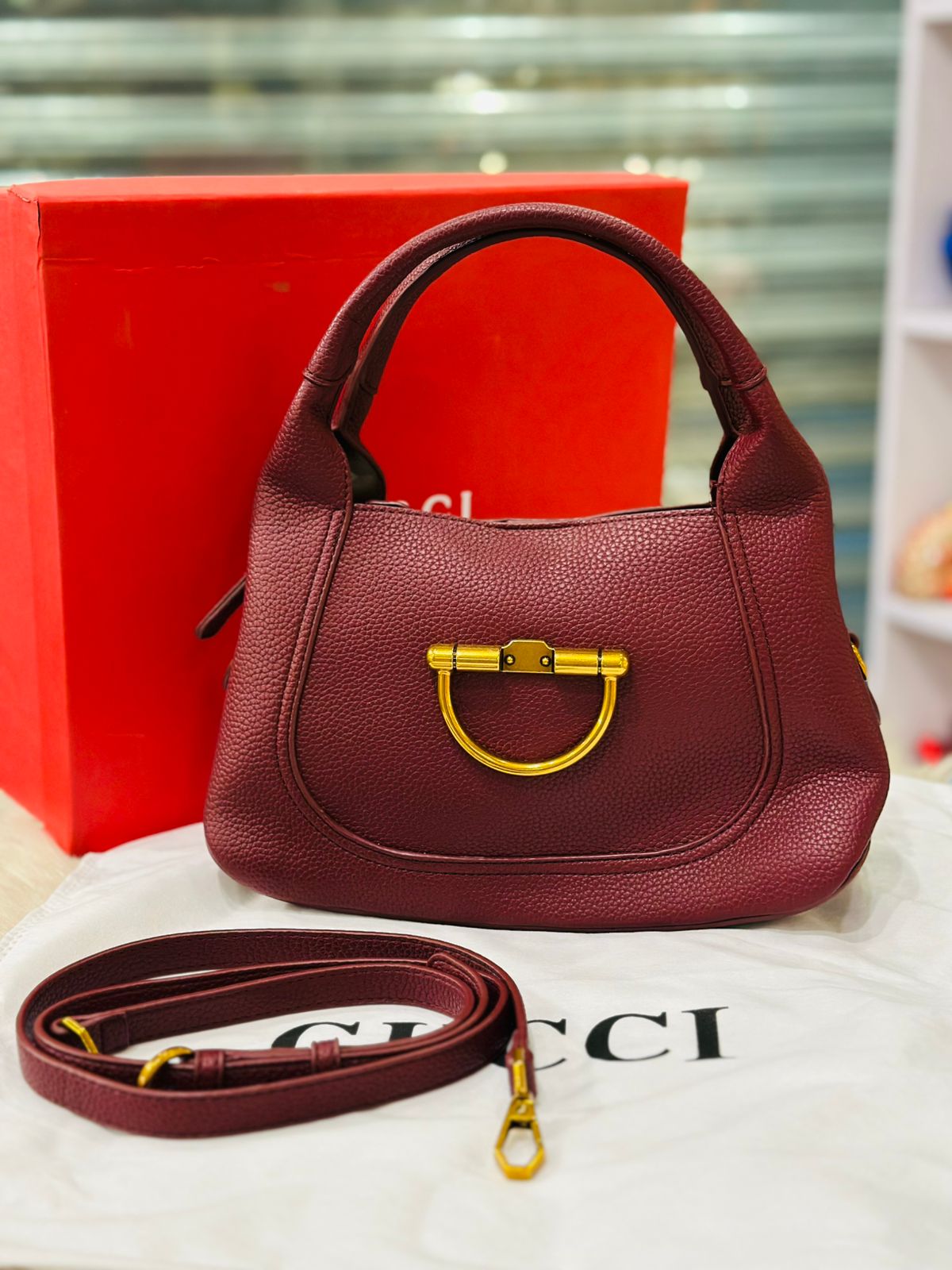 Maroon handbag with gold hardware on a Gucci branded surface, with a red box in the background.
